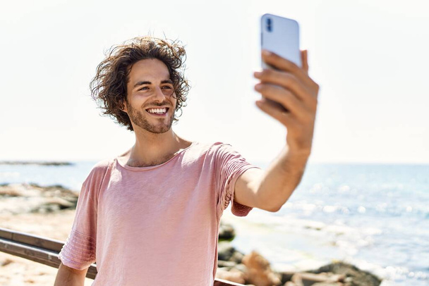 Νεαρός Ισπανός χαμογελώντας χαρούμενος βγάζει σέλφι δίπλα στο smartphone που στέκεται στην παραλία.. - Φωτογραφία, εικόνα
