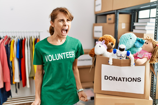 Beautiful caucasian woman wearing volunteer t shirt at donations stand angry and mad screaming frustrated and furious, shouting with anger. rage and aggressive concept.  - Photo, Image