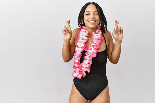 Young african american woman wearing swimsuit and hawaiian lei gesturing finger crossed smiling with hope and eyes closed. luck and superstitious concept.  - Фото, изображение