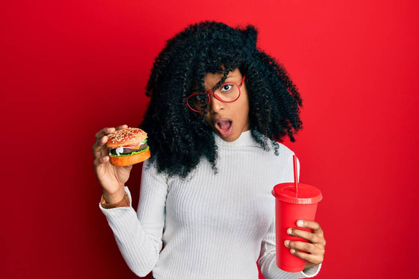Αφροαμερικανή γυναίκα με αφρο μαλλιά τρώει ένα νόστιμο κλασικό burger και σόδα στο πρόσωπο σοκ, αναζητούν δύσπιστοι και σαρκαστική, έκπληκτος με ανοιχτό στόμα  - Φωτογραφία, εικόνα