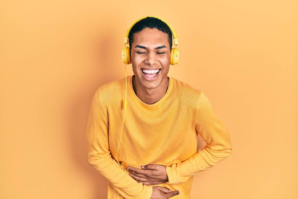 Young african american guy listening to music using headphones smiling and laughing hard out loud because funny crazy joke with hands on body.  - Фото, изображение