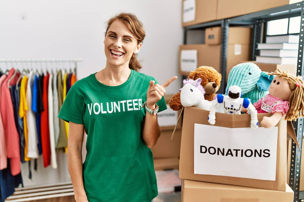 Beautiful caucasian woman wearing volunteer t shirt at donations stand cheerful with a smile on face pointing with hand and finger up to the side with happy and natural expression  - Photo, Image