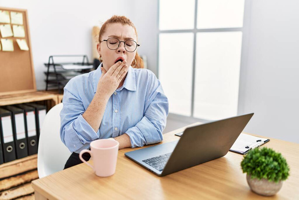 Молодая рыжая женщина, работающая в офисе с помощью ноутбука, скучала зевая усталая, прикрывая рот рукой. беспокойство и сонливость.  - Фото, изображение