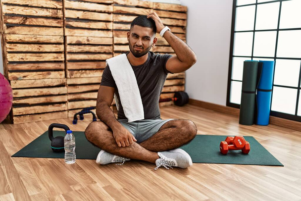 Молодой индиец, сидящий на тренировочном коврике в спортзале, смущается и задается вопросом. неуверенный в сомнении, думающий с рукой на голове. застенчивое понятие.  - Фото, изображение