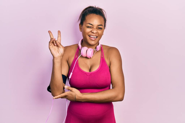 Mladá africká americká dívka v tělocvičně oblečení a pomocí sluchátek s úsměvem s veselou tváří mrkající na kameru dělat vítězství znamení s prsty. číslo dvě.  - Fotografie, Obrázek