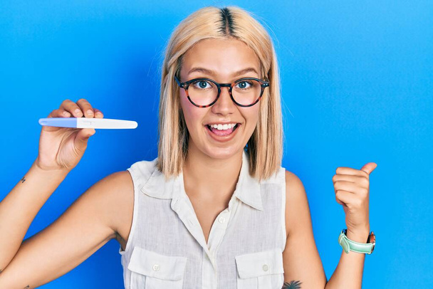 Beautiful blonde woman holding pregnancy test result pointing thumb up to the side smiling happy with open mouth  - Φωτογραφία, εικόνα
