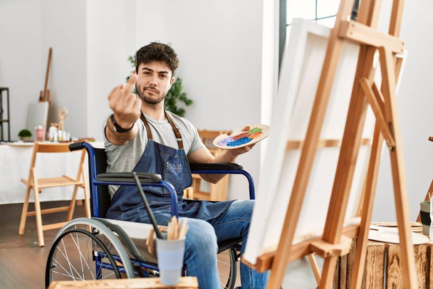 Young hispanic man sitting on wheelchair painting at art studio showing middle finger, impolite and rude fuck off expression  - Fotoğraf, Görsel