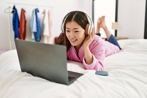 Joven chica china usando el ordenador portátil y auriculares acostados en la cama en el dormitorio. - Foto, Imagen