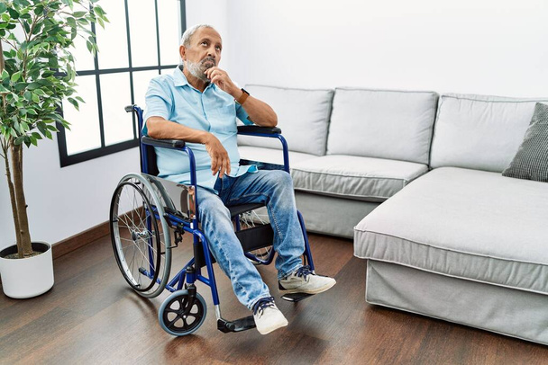 Pohledný starší muž sedící na vozíčku v obývacím pokoji s rukou na bradě a přemýšlející o otázce a zamyšleném výrazu. s zamyšleným úsměvem. pochybný koncept.  - Fotografie, Obrázek