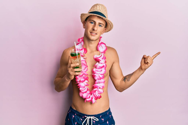Junger kaukasischer Mann mit Sommermütze und hawaiianischem Lei, der einen Cocktail trinkt, lächelt glücklich und zeigt mit Hand und Finger zur Seite  - Foto, Bild