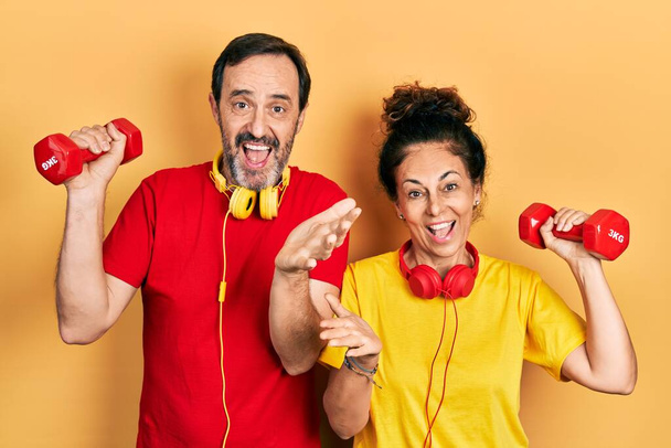 Para Hiszpanek w średnim wieku i mężczyzna ubrani w odzież sportową z hantlami świętującymi osiągnięcie ze szczęśliwym uśmiechem i wyrazem zwycięzcy z podniesioną ręką  - Zdjęcie, obraz
