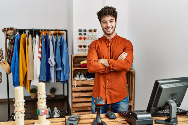 Młody latynoski sklepikarz uśmiecha się szczęśliwy stojąc z rękoma skrzyżowanymi w sklepie odzieżowym. - Zdjęcie, obraz