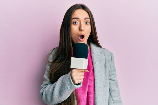 Young hispanic girl holding reporter microphone scared and amazed with open mouth for surprise, disbelief face  - Fotoğraf, Görsel