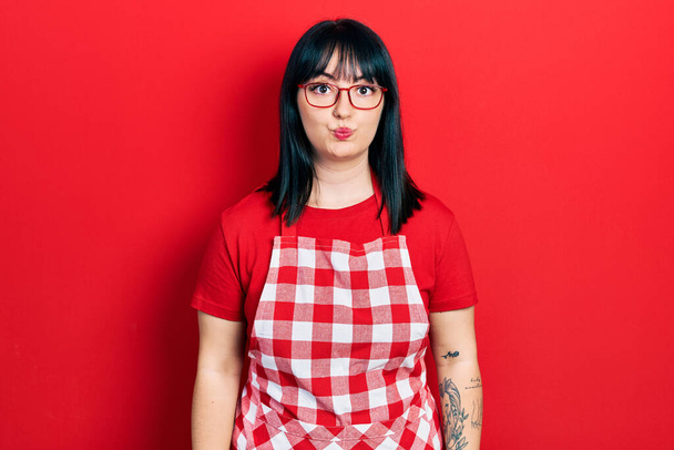 Young hispanic woman wearing cook apron and glasses puffing cheeks with funny face. mouth inflated with air, crazy expression.  - Фото, изображение