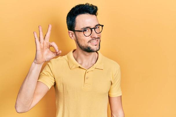 Junger hispanischer Mann in lässiger Kleidung und Brille, der positiv lächelt und mit Händen und Fingern Okay-Zeichen macht. Erfolgreicher Ausdruck.  - Foto, Bild
