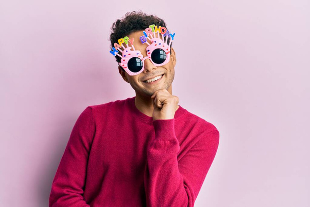 Handsome hispanic man wearing funny happy birthday glasses smiling looking confident at the camera with crossed arms and hand on chin. thinking positive.  - Фото, изображение