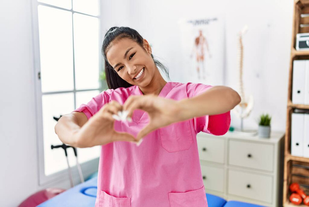 Молода іспаномовна жінка, яка працює в клініці відновлення болю, посміхається в коханні, роблячи символ серця формою руками. романтична концепція
.  - Фото, зображення