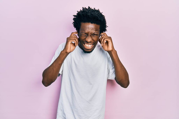 Giovane uomo afroamericano indossa casual t shirt bianca che copre le orecchie con le dita con espressione infastidita per il rumore della musica ad alto volume. concetto di sordo.  - Foto, immagini