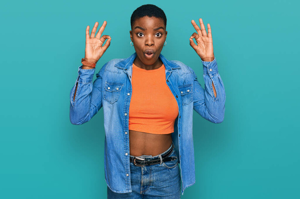 Joven mujer afroamericana vistiendo ropa casual que se ve sorprendida y sorprendida haciendo símbolo de aprobación aceptable con los dedos. expresión loca  - Foto, Imagen