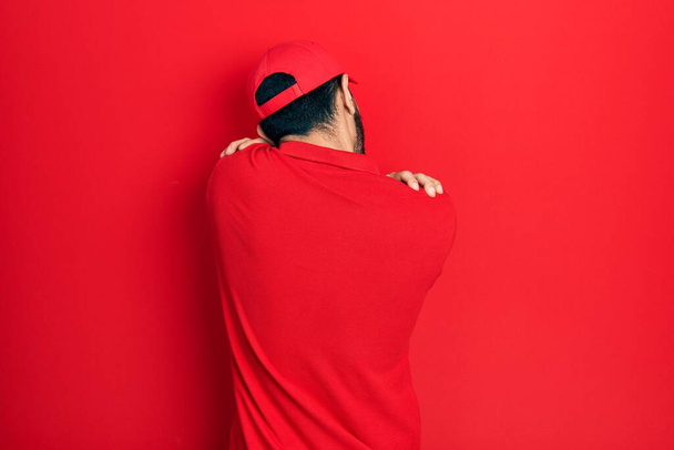 Hispanic man with beard wearing delivery uniform and cap hugging oneself happy and positive from backwards. self love and self care  - Photo, Image