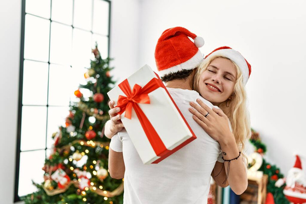 Young couple wearing christmas hat hugging and holding gift at home. - Photo, Image
