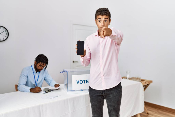 Jonge Latijns-Amerikaanse mannen bij de politieke campagne verkiezing houden smartphone wijzend met de vinger naar de camera en naar u, zelfverzekerd gebaar kijken serieus  - Foto, afbeelding