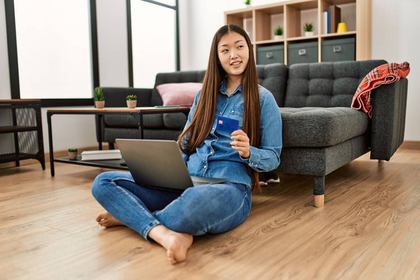 Młoda chińska dziewczyna za pomocą laptopa i karty kredytowej siedzi na podłodze w domu. - Zdjęcie, obraz