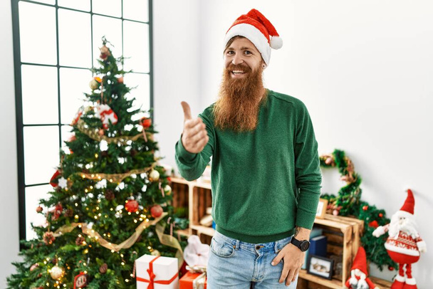 Redhead man with long beard wearing christmas hat by christmas tree smiling friendly offering handshake as greeting and welcoming. successful business.  - Fotoğraf, Görsel