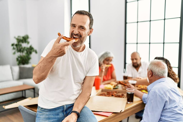 Група людей середнього віку посміхаються щасливо їдять італійську піцу, сидячи на столі вдома
 - Фото, зображення