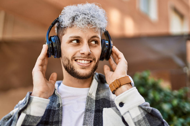 Joven hombre hispano sonriendo feliz usando auriculares en la ciudad. - Foto, imagen