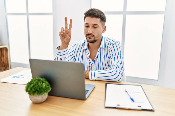 Young handsome man with beard working at the office using computer laptop showing and pointing up with fingers number two while smiling confident and happy.  - Foto, Bild