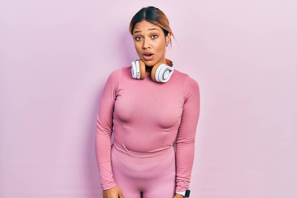Beautiful hispanic woman wearing gym clothes and using headphones in shock face, looking skeptical and sarcastic, surprised with open mouth  - Foto, imagen