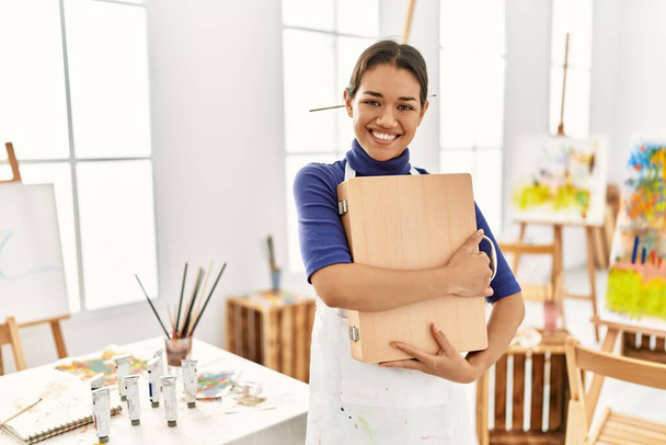 Young latin woman smiling confident hugging draw briefcase at art studio - Photo, Image