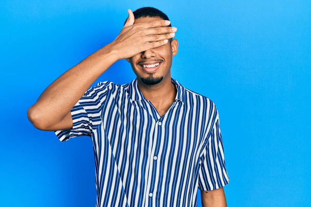 Mladý africký Američan, oblečený do neformálního oblečení, se usmívá a směje se s rukou na tváři, zakrývající oči, aby ho překvapil. slepý koncept.  - Fotografie, Obrázek
