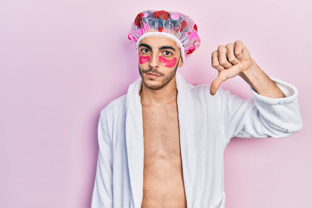 Jonge Latijns-Amerikaanse man met badjas en douchepet met behulp van oogzakken patches met boos gezicht, negatief teken toont een hekel aan met duimen naar beneden, afwijzing concept  - Foto, afbeelding