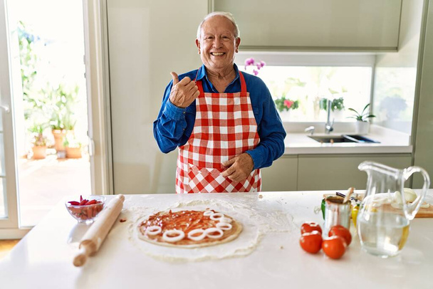 Hombre mayor con pelo gris cocinando pizza en la cocina casera sonriendo con la cara feliz mirando y señalando a un lado con el pulgar hacia arriba.  - Foto, Imagen