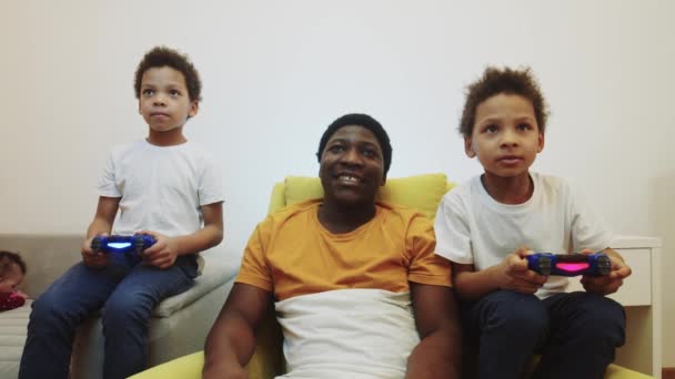 Kaksi pientä mustaa poikaa pelaa peliä joysticksin avulla ja heidän isänsä istuu niiden välissä. - Materiaali, video