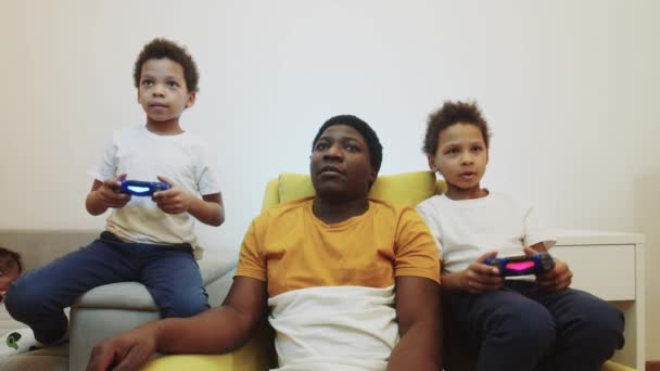 Δύο μικρά μαύρα αγόρια παίζουν ένα παιχνίδι χρησιμοποιώντας joysticks και ο πατέρας τους κάθεται ανάμεσά τους και να βοηθήσει - Πλάνα, βίντεο