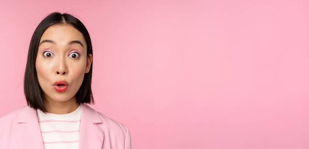 Close up portrait of asian corporate woman, business lady looking surprised and amazed at camera, standing in suit against pink background - Foto, Imagem