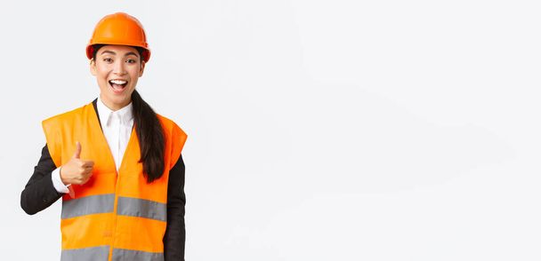 Υποστηρικτική και ευχαριστημένη, ευτυχισμένη Ασιάτισσα αρχιμηχανικός με κράνος ασφαλείας, που δείχνει τους αντίχειρες πάνω στην έγκριση, ικανοποιημένη με τις κατασκευαστικές εργασίες. Βιομηχανική γυναίκα σε reflecive σακάκι επιθεωρήσει περιοχή - Φωτογραφία, εικόνα