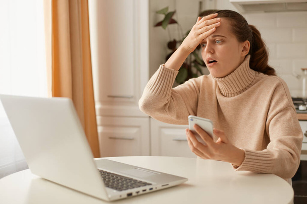 Indoor shot of astonished scared woman wearing beige sweater posing in kitchen, looking at cell phone screen with open mouth, touching head, forgot important information, mistake. - Photo, Image