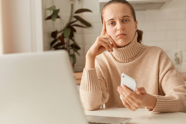 Indoor shot of sad pensive woman wearing beige sweater posing in kitchen, holding smart phone and looking at camera with thoughtful facial expression. - Photo, Image