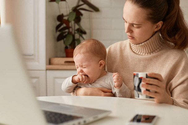 Indoor shot of sad dark haired woman wearing beige sweater posing in kitchen with her crying baby and holding cup of tea or coffee, trying to calm down kid. - Фото, зображення
