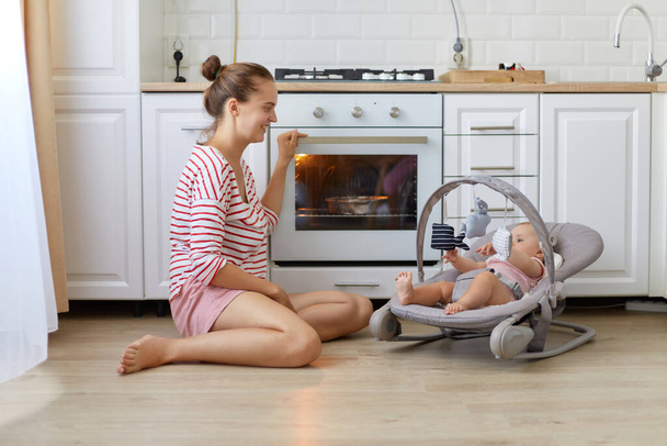 Indoor shot of mother baking a pie with her newborn daughter lying in rocking chair, woman wearing casual style shirt, female cooking in kitchen, baking pastry. - Φωτογραφία, εικόνα