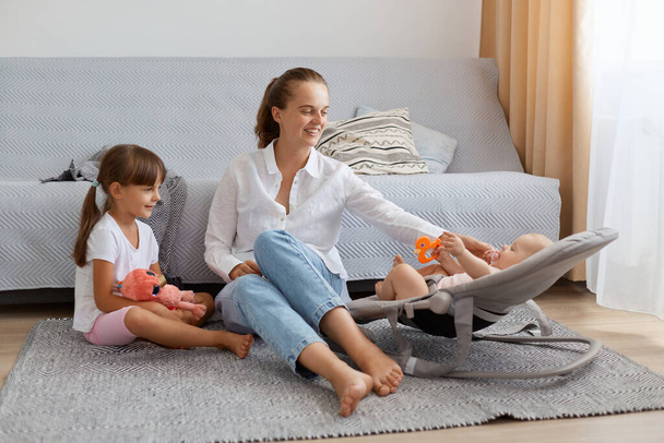 Portrait of smiling woman with ponytail wearing white shirt and jeans sitting on floor with her daughters, playing with toddler kid in rocking chair together with elder child. - Foto, Bild