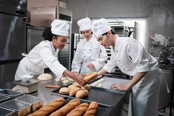 Profesionální gurmánský tým, tři mladí kuchaři v bílých kuchařských uniformách a zástěrách hnětou těsto a vejce, připravují chléb a čerstvé pečivo, pečou v troubě v kuchyni restaurace z nerezové oceli. - Fotografie, Obrázek