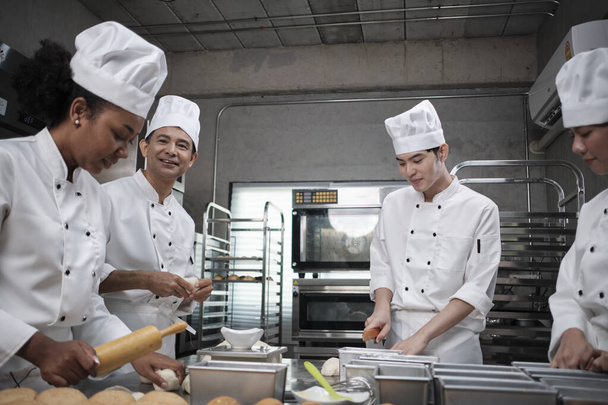 Multiracial professional gourmet team, four chefs in white cook uniforms and aprons knead pastry dough and flour, prepare bread, and bakery food, baking in oven at stainless steel restaurant kitchen. - Valokuva, kuva