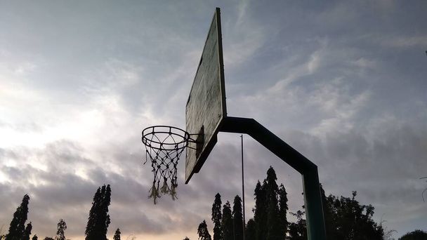 Πρόσθια όψη γωνίας αμυδρό πράσινο παλιό στεφάνι μπάσκετ και σπασμένο δίχτυ με μπλε φόντο του πρωινού ουρανού στο πεδίο του δημόσιου αθλητισμού. - Φωτογραφία, εικόνα