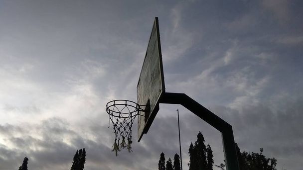 Voorste hoek uitzicht op dimgroene oude basketbal hoepel en gebroken net met een blauwe achtergrond van de ochtend hemel in het publieke sportveld. - Foto, afbeelding