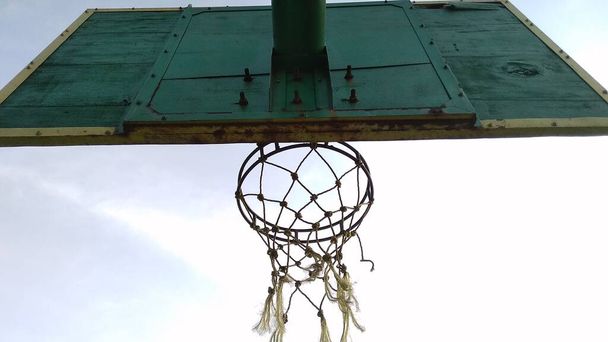 薄緑色の古いバスケットボールのフープと公共のスポーツフィールドで朝の空の青の背景と壊れたネットのフロントバック角度ビュー. - 写真・画像